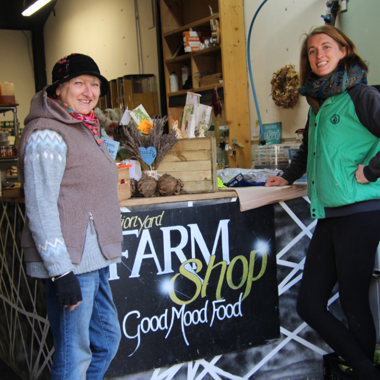Two women pose in a farm shop, smiling and standing behind a counter with produce.
